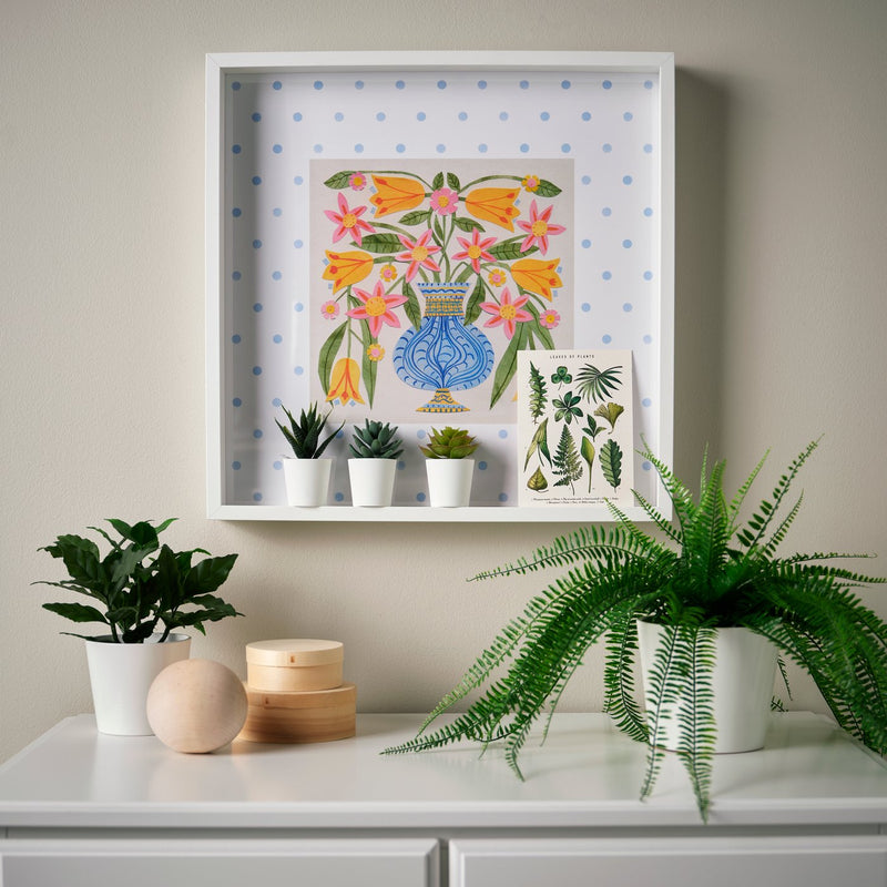 Una cornice bianca SANNAHED con un poster di un vaso blu con dei fiori. Sotto un mobile bianco con piante in vaso.
