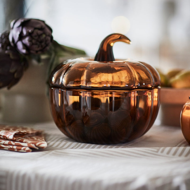 Primo piano di una ciotola decorativa con coperchio HÖSTAGILLE color ambra a forma di zucca su un tavolo con una tovaglia fantasia.
