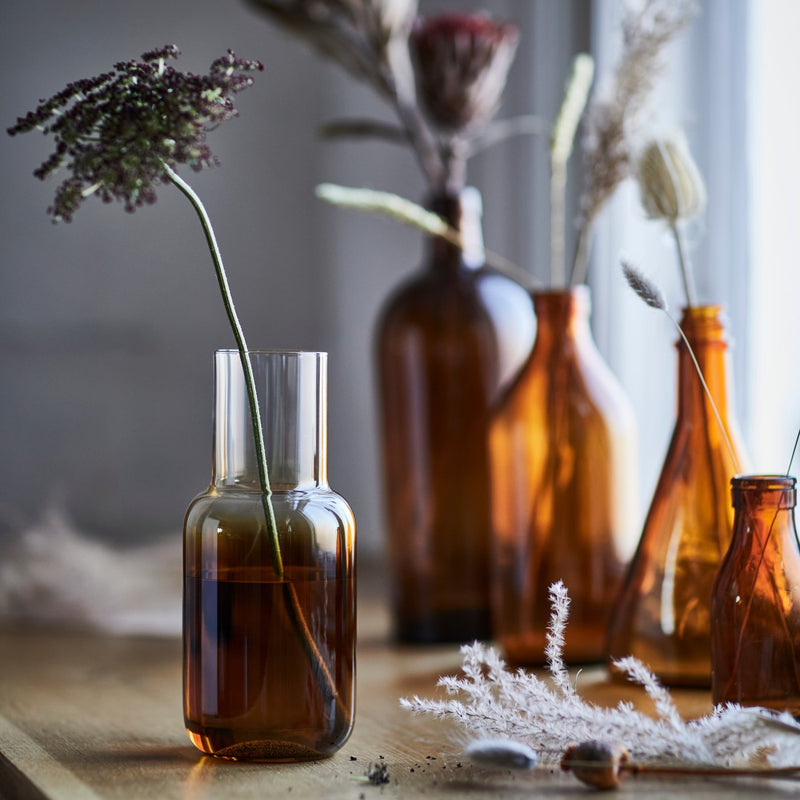 Un vaso HÖSTAGILLE marrone con un fiore essiccato, piacevolmente appoggiato su un tavolo in legno illuminato da una luce d'atmosfera soffusa.